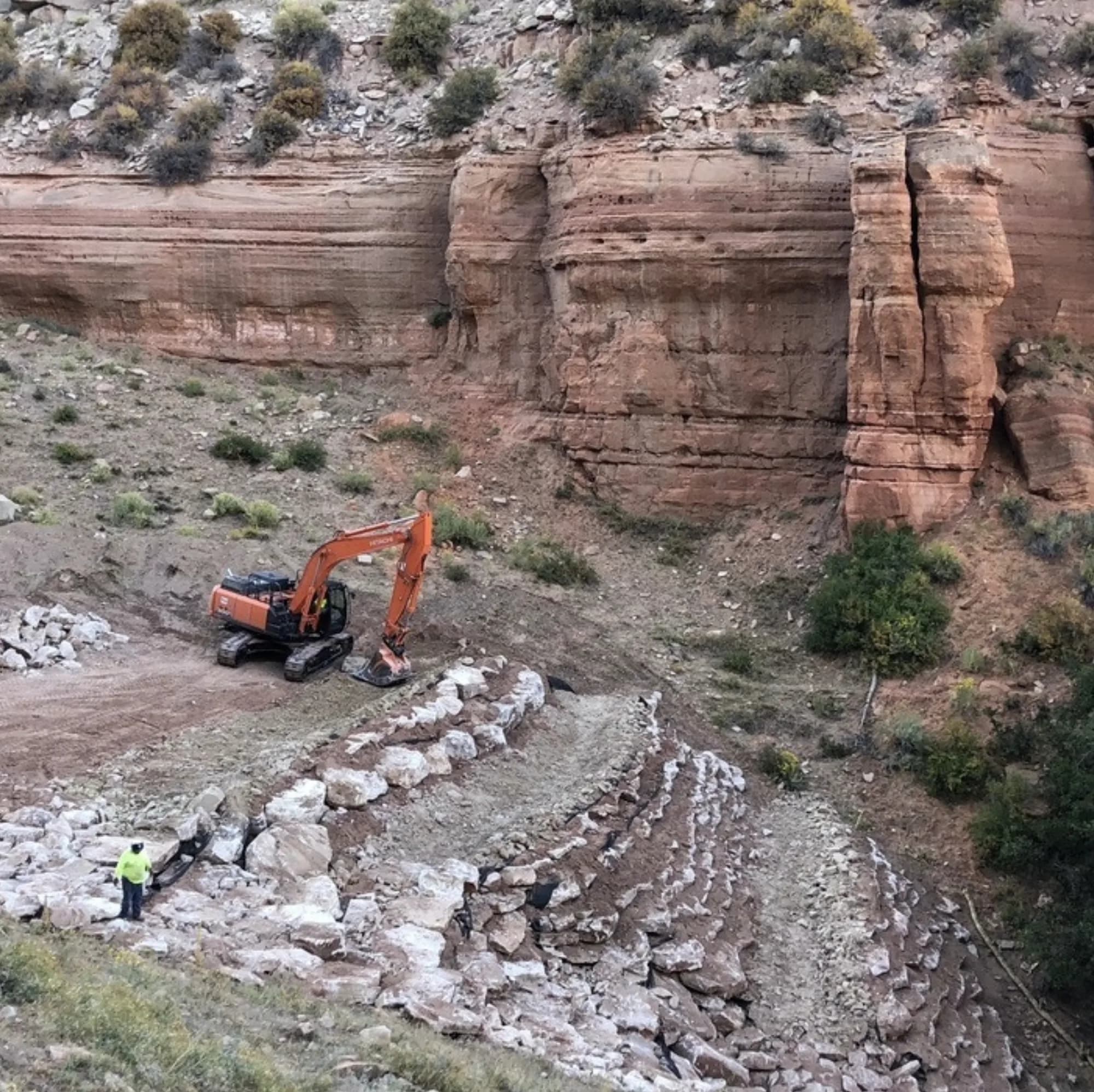 Excavator and crew placing rock armoring at the base of red canyon cliffs — Clawson Excavating remote site operations
